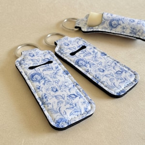 Puede incluir: Tres fundas rectangulares para bálsamo labial con llaveros, con un exterior de tela blanca con estampado floral azul. Cada funda tiene un borde negro y una abertura de ranura horizontal. Una funda contiene un tubo de bálsamo labial blanco.