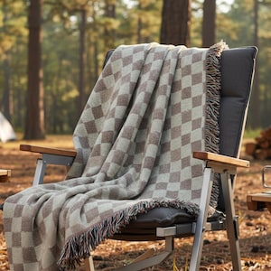 May include: A brown and beige checkered blanket with fringe is draped over a folding camping chair. The chair has a gray seat and back with wooden armrests. The blanket's pattern is a repeating diamond design.