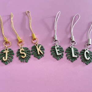 May include: Six personalized phone charms with a tropical leaf design. Each charm features a dark green leaf with a gold or silver initial: J, S, K, E, L, and C. The charms have matching gold or silver cords and are set against a pink background.