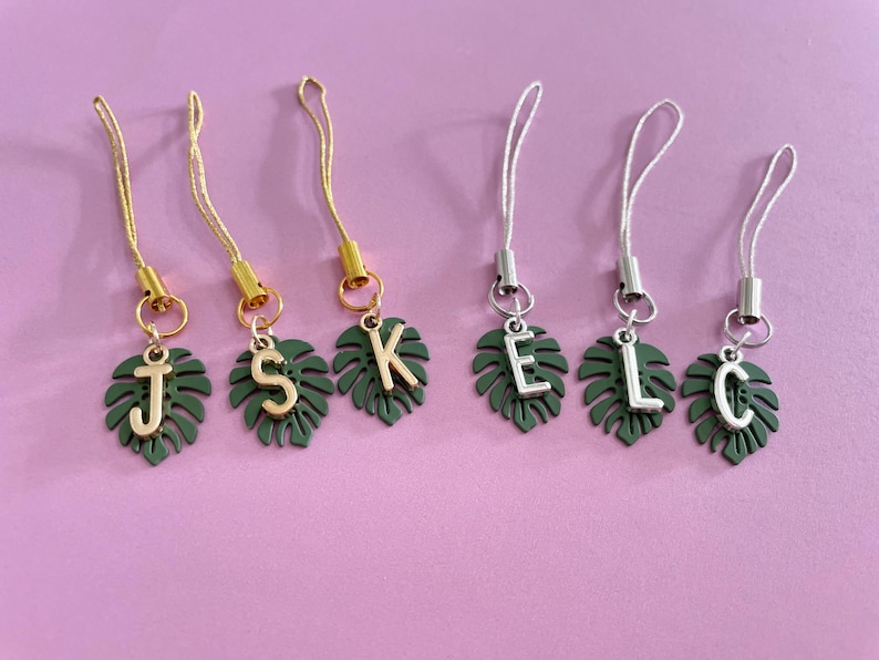 May include: Six personalized phone charms with a tropical leaf design. Each charm features a dark green leaf with a gold or silver initial: J, S, K, E, L, and C. The charms have matching gold or silver cords and are set against a pink background.