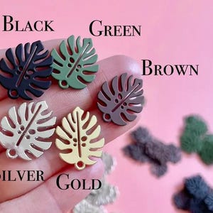 May include: Close-up of several monstera leaf charms in various colors: black, green, brown, silver, and gold. The word "BLACK", "GREEN", "BROWN", "SILVER", and "GOLD" are written above the charms. The charms are held in a hand.