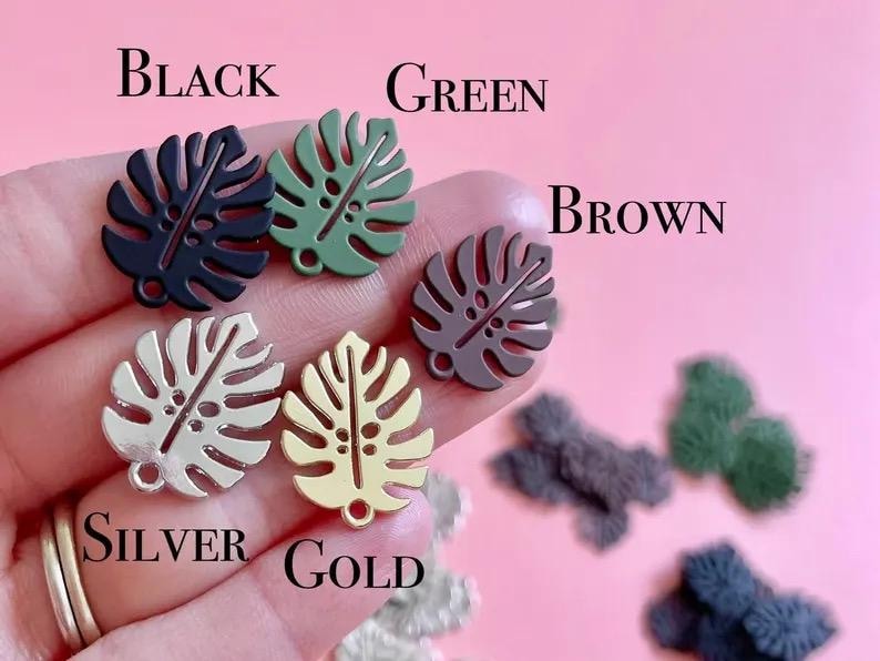 May include: Close-up of several monstera leaf charms in various colors: black, green, brown, silver, and gold. The word "BLACK", "GREEN", "BROWN", "SILVER", and "GOLD" are written above the charms. The charms are held in a hand.