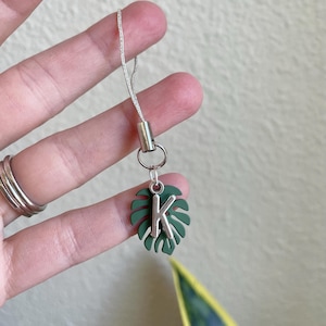 May include: A silver-toned charm with a green monstera leaf design and the letter "K". The charm is attached to a silver cord. The charm is held up by a hand.