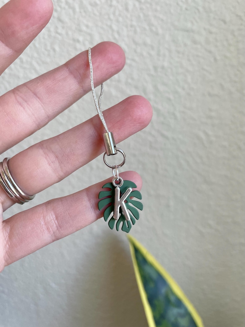 May include: A silver-toned charm with a green monstera leaf design and the letter "K". The charm is attached to a silver cord. The charm is held up by a hand.