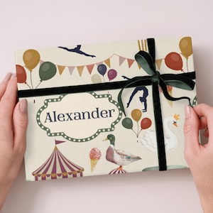 May include: A gift box with a circus theme, decorated with balloons, a circus tent, and a swan. The box is tied with a black velvet ribbon and has the name "Alexander" on the front.