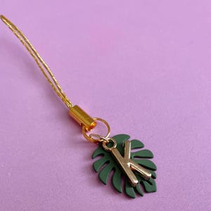 May include: A gold-colored charm with a green leaf and the letter "K" in gold. The charm is attached to a gold-colored cord. The background is a solid light purple color.