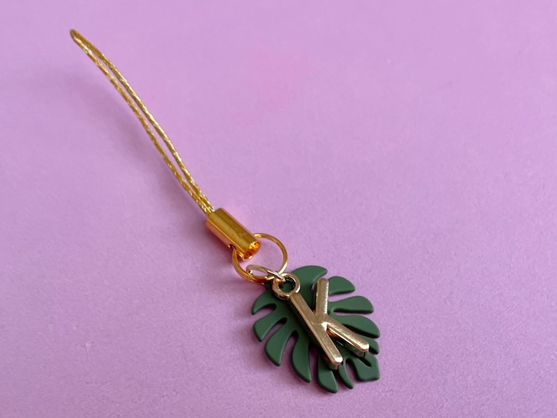 May include: A gold-colored charm with a green leaf and the letter "K" in gold. The charm is attached to a gold-colored cord. The background is a solid light purple color.