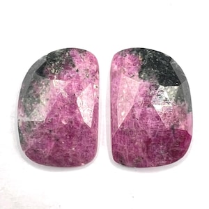 Peut inclure: Deux pierres précieuses facettées en forme de bouclier, présentant une couleur rose magenta vibrante mélangée à des inclusions gris anthracite foncé. Les pierres ont une surface polie et réfléchissante avec de multiples facettes géométriques.