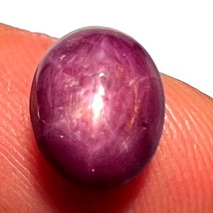 Peut inclure: Gros plan sur une pierre précieuse polie, de forme ovale, rouge-violacé. La gemme a une surface lisse avec des motifs internes subtils et un point brillant et réfléchissant. La pierre est tenue par un doigt.