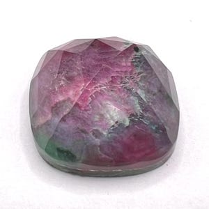 May include: A faceted, cushion-shaped gemstone displaying a mix of deep magenta, purple, and dark green hues. The surface features a geometric cut with multiple triangular facets that reflect light, showcasing internal natural inclusions and color variations.