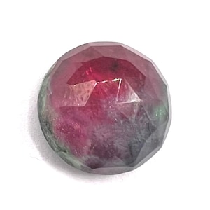 May include: A round, faceted gemstone displaying a gradient of deep magenta and pink hues transitioning into dark green and charcoal gray tones at the edges. The stone features a polished, multi-faceted surface that reflects light across its circular shape.