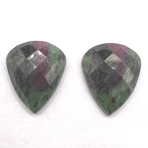 May include: Two pear-shaped ruby zoisite gemstones featuring a faceted surface. The stones display a mix of deep green and magenta-red colors with natural inclusions visible throughout the polished, geometric cut of each individual piece.