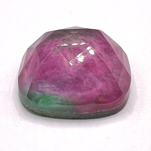 May include: A faceted, dome-shaped gemstone featuring a vibrant mix of deep magenta and bright green colors. The stone has a smooth, polished surface with multiple geometric facets that reflect light across its rounded, cabochon-like top profile.