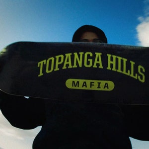 Könnte beinhalten: Eine Person hält ein schwarzes Skateboard-Deck mit gelber Aufschrift "Topanga Hills Mafia".