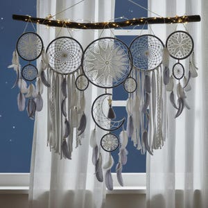 May include: A handcrafted dreamcatcher with multiple circular and crescent moon designs. The dreamcatchers are white and gray, adorned with feathers and hanging from a wooden branch with fairy lights. The background is a blue wall and white curtains.