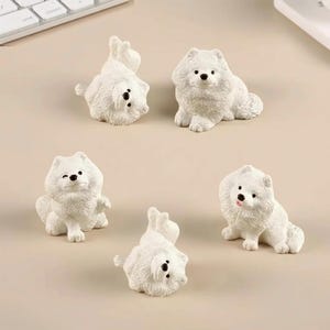 Puede incluir: Un conjunto de cinco pequeñas figuras de perro blancas, cada una con pelaje esponjoso, orejas puntiagudas y nariz negra. Las figuras se muestran en varias posturas, incluyendo sentadas, tumbadas y una guiñando un ojo.
