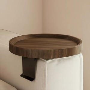 Puede incluir: Una bandeja redonda de madera marrón oscuro para el brazo del sofá con un borde elevado. La bandeja está sujeta a un soporte de metal negro que descansa sobre el brazo de un sofá blanco. Ofrece una superficie práctica para bebidas o aperitivos.