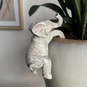 May include: A white elephant figurine climbing on the edge of a ceramic pot. The elephant has a textured surface and is positioned as if it's hanging on. The pot is light gray with a brown rim, and green plant leaves are visible.
