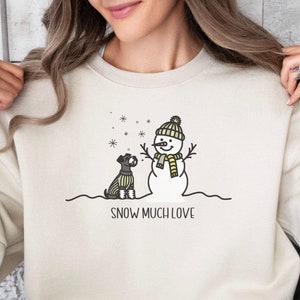 May include: A cream colored sweatshirt with a graphic of a snowman and a dog wearing a scarf. The text "SNOW MUCH LOVE" is printed below the graphic.