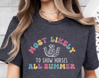Könnte beinhalten: Ein dunkelgraues T-Shirt mit dem Text "Most Likely to Show Horses All Summer" in Regenbogenfarben. Der Text ist in einem Bogen angeordnet, in der Mitte befindet sich ein Hufeisen-Motiv.