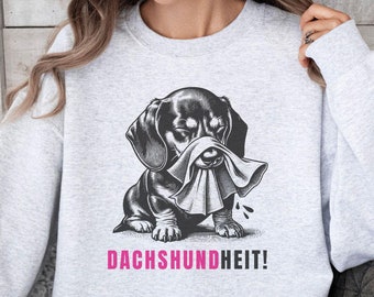 Op de afbeelding: Lichtgrijs sweatshirt met een zwart-wit illustratie van een teckel die zijn neus snuit met een tissue. De roze tekst "DACHSHUNDHEIT!" is eronder gedrukt. Het sweatshirt heeft een casual stijl.