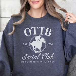 May include: A dark gray sweatshirt with a white graphic design that says "OTTB Social Club Est. 1730 We do more than just run". The graphic features a silhouette of a jockey riding a horse.