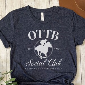 May include: A dark blue t-shirt with a white graphic design that says "OTTB Social Club Est. 1730 We do more than just run". The graphic also features a silhouette of a jockey riding a horse.