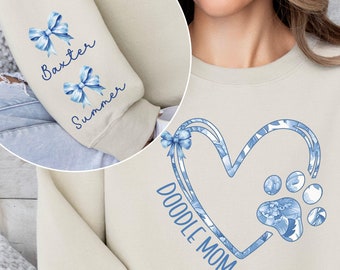 May include: A beige sweatshirt featuring a blue floral heart design with the text "Doodle Mom". The sleeves have blue bows with the names "Baxter" and "Summer" written on them.