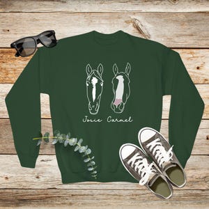 May include: A dark green sweatshirt with a white outline of two horse heads and the names "Josie" and "Carmel" printed on the front.