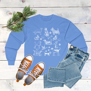 May include: A light blue long-sleeved t-shirt with a white graphic print of various dog breeds. The graphic includes a variety of dog breeds, including a dachshund, a poodle, a terrier, and a retriever. The shirt is a casual and comfortable style.
