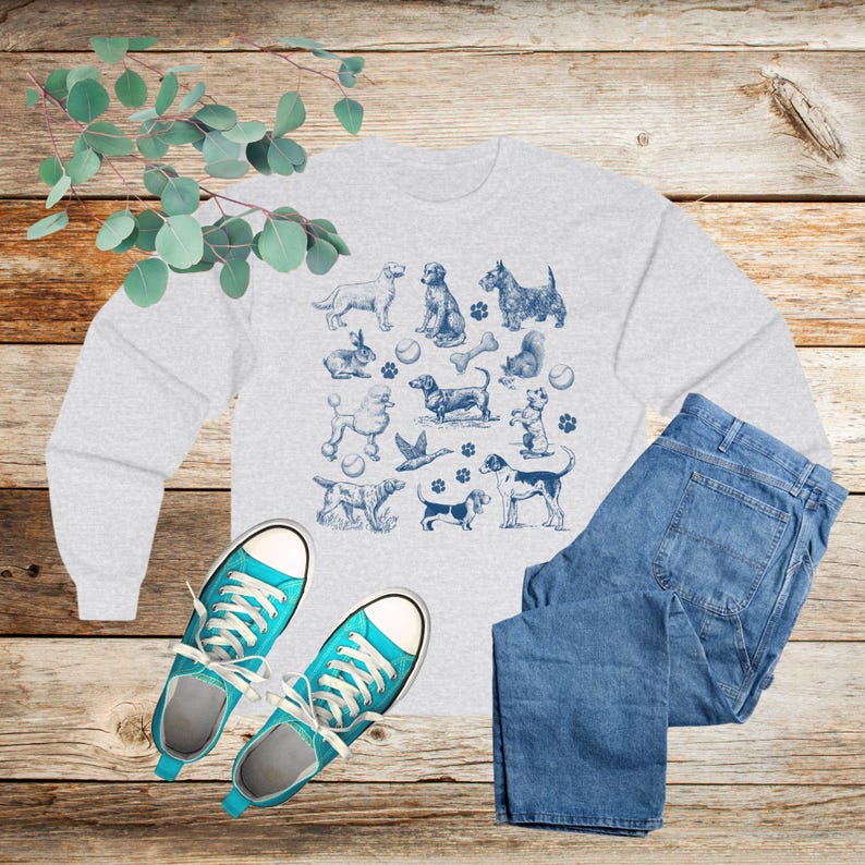 May include: A gray sweatshirt with a blue print of various dog breeds, bones, and balls. The sweatshirt is laid out on a wooden surface with a pair of blue jeans and blue sneakers.
