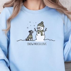 May include: A light blue sweatshirt with a snowman and a small dog in front of it. The snowman is wearing a yellow and white hat and scarf. The text "SNOW MUCH LOVE" is printed below the snowman and dog.