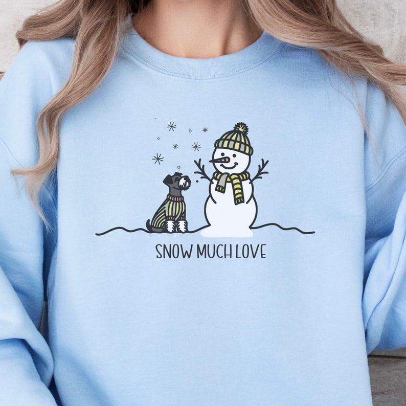May include: A light blue sweatshirt with a snowman and a small dog in front of it. The snowman is wearing a yellow and white hat and scarf. The text "SNOW MUCH LOVE" is printed below the snowman and dog.