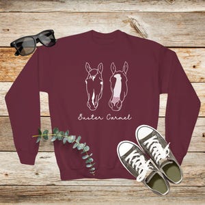 May include: Burgundy sweatshirt with a white outline of two horse heads and the text "Buster Carmel" printed on the front.