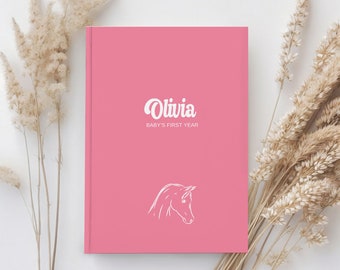May include: Pink baby journal with the name Olivia and the text "Baby's First Year" in white. A white horse silhouette is in the bottom center of the cover.