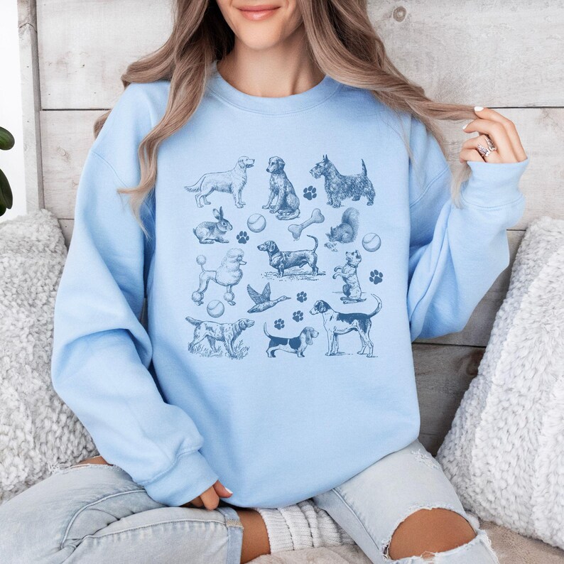 Peut inclure: Un sweat-shirt bleu clair avec un imprim&eacute; graphique de diff&eacute;rentes races de chiens, dont un caniche, un teckel et un terrier &eacute;cossais. L'imprim&eacute; comprend &eacute;galement des os de chien, des balles et des empreintes de pattes.