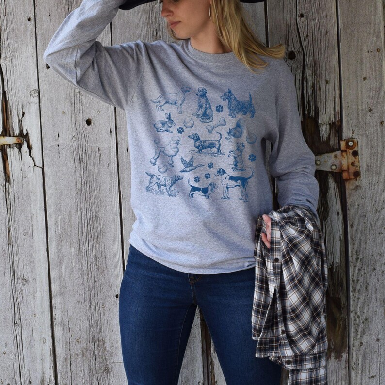 May include: A gray long-sleeved sweatshirt with a blue print of various dog breeds. The print includes a dachshund, a poodle, a terrier, and a retriever. The sweatshirt is being worn by a person with long blonde hair.