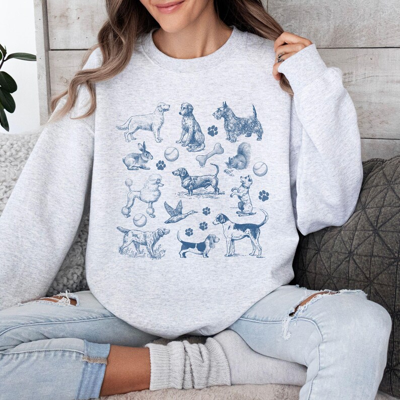 Peut inclure: Un sweat-shirt gris avec un imprim&eacute; bleu de diff&eacute;rentes races de chiens, dont un caniche, un teckel, un basset hound et un beagle. L'impression comprend &eacute;galement des os, des balles et d'autres articles li&eacute;s aux chiens.