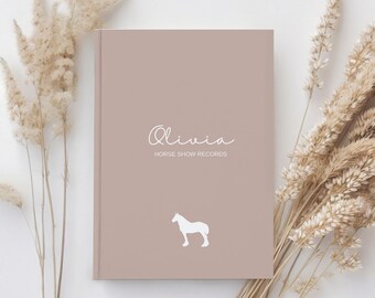 Può includere: Un quaderno marrone chiaro con il nome "Olivia" e il testo "Horse Show Records" stampato sulla copertina. Sulla copertina è anche stampata una silhouette bianca di un cavallo.