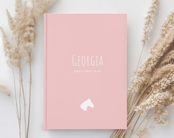 May include: A pink baby journal with the name "GEORGIA" and the text "BABY'S FIRST YEAR" on the front cover. A white horse silhouette is in the bottom center of the cover.
