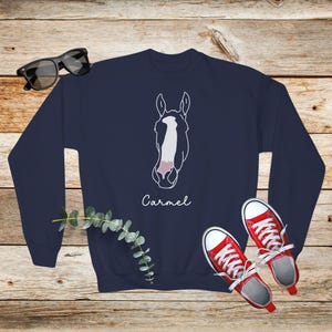 May include: Navy blue sweatshirt with a white outline of a horse's head and the name "Carmel" printed below. The sweatshirt is laying on a wooden surface with a pair of red sneakers and a sprig of greenery.
