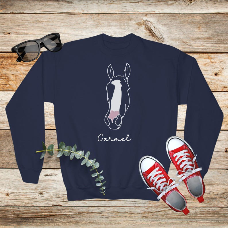 May include: Navy blue sweatshirt with a white outline of a horse's head and the name "Carmel" printed below. The sweatshirt is laying on a wooden surface with a pair of red sneakers and a sprig of greenery.