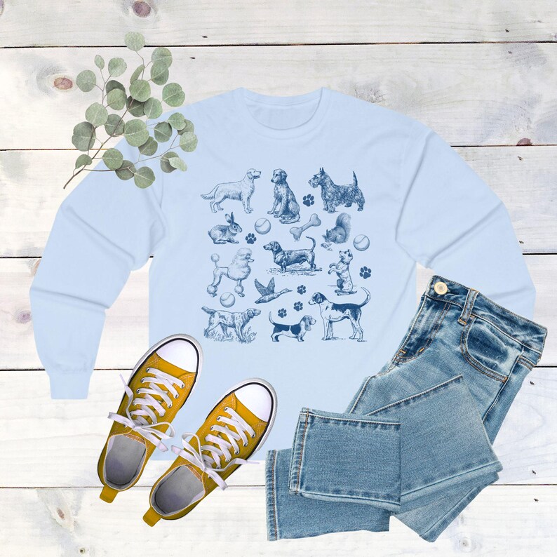 May include: Light blue long-sleeved sweatshirt with a graphic print of various dogs and dog-related items, including bones, balls, and paw prints.