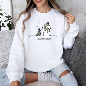 May include: White sweatshirt with a snowman and a dog wearing a scarf. The text "SNOW MUCH LOVE" is printed below the image.