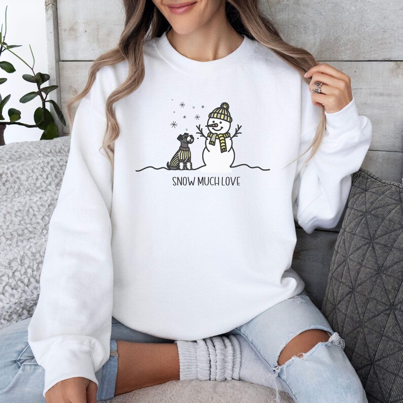 May include: White sweatshirt with a snowman and a dog wearing a scarf. The text "SNOW MUCH LOVE" is printed below the image.