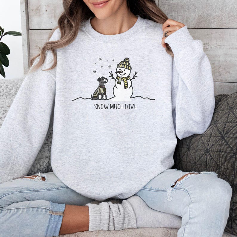 May include: A gray sweatshirt with a snowman and a small dog wearing a scarf. The text "SNOW MUCH LOVE" is printed below the image.
