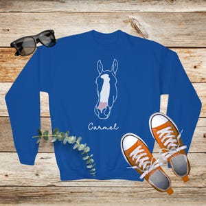 May include: A royal blue sweatshirt with a white outline of a horse's head and the name "Carmel" printed below. The sweatshirt is laying on a wooden surface with a pair of orange sneakers.