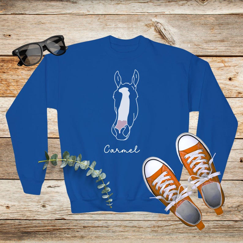 May include: A royal blue sweatshirt with a white outline of a horse's head and the name "Carmel" printed below. The sweatshirt is laying on a wooden surface with a pair of orange sneakers.