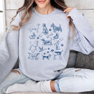 Peut inclure: Un sweat-shirt gris avec un imprim&eacute; bleu de diff&eacute;rentes races de chiens, des os et des balles de tennis. L'impression est une illustration de style vintage.