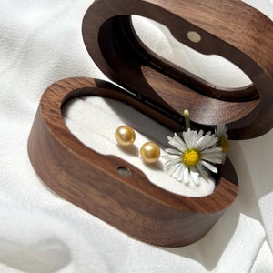 May include: Two golden pearl earrings in a wooden box with a white daisy.